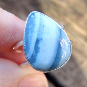 May include: A teardrop-shaped ring featuring a large, light blue gemstone with white and darker blue veining. The stone is set in a silver-colored metal band. The ring's design is simple, with a focus on the natural beauty of the stone.