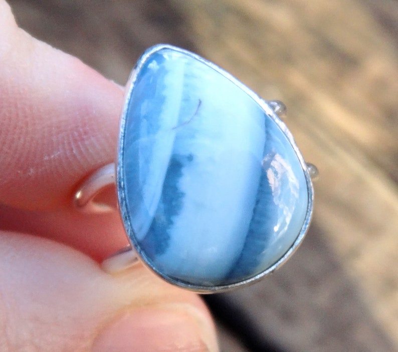 May include: A teardrop-shaped ring featuring a large, light blue gemstone with white and darker blue veining. The stone is set in a silver-colored metal band. The ring's design is simple, with a focus on the natural beauty of the stone.