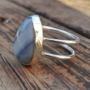 May include: A silver ring with a large, teardrop-shaped, blue-grey stone. The ring has a double-band design and a gold-colored bezel setting. The stone's surface has subtle, natural patterns. The ring is displayed on a wooden surface.