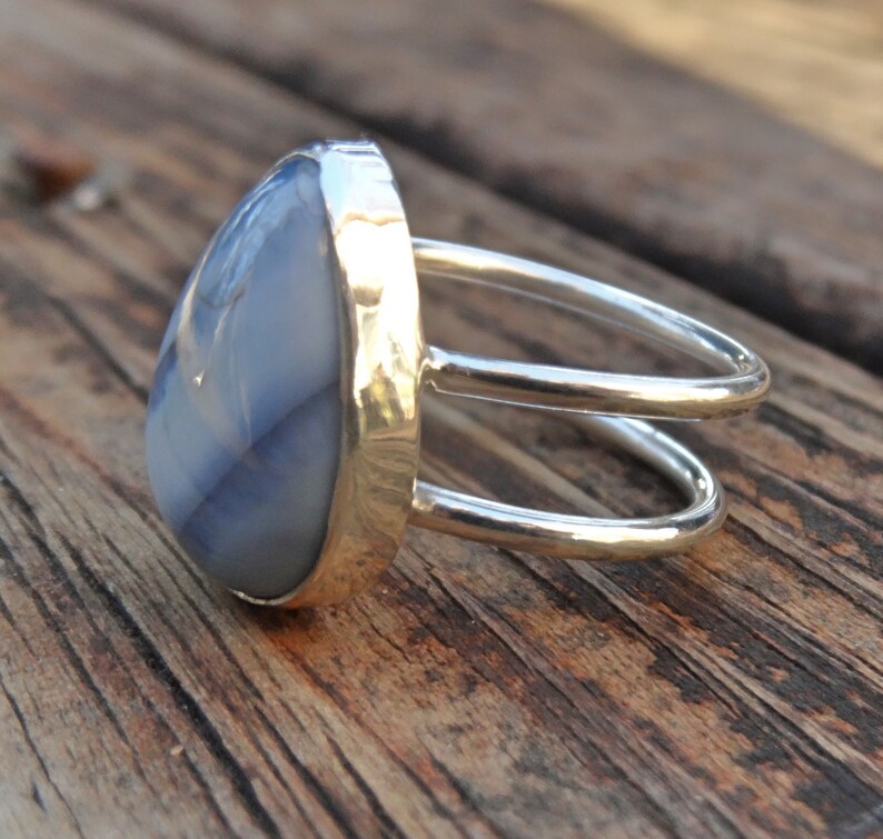 May include: A silver ring with a large, teardrop-shaped, blue-grey stone. The ring has a double-band design and a gold-colored bezel setting. The stone's surface has subtle, natural patterns. The ring is displayed on a wooden surface.