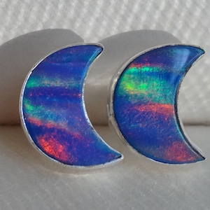 May include: A pair of silver crescent moon earrings with iridescent blue opal stones. The stones have a rainbow of colors, including green, blue, and red.