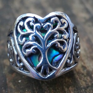 May include: A silver ring with a heart-shaped design featuring a tree of life with abalone inlay. The ring has intricate details and a Celtic knot design on the band.