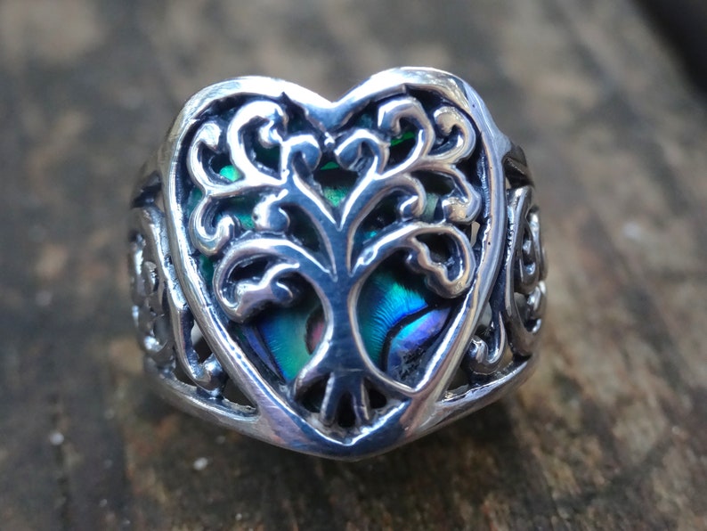 May include: A silver ring with a heart-shaped design featuring a tree of life with abalone inlay. The ring has intricate details and a Celtic knot design on the band.