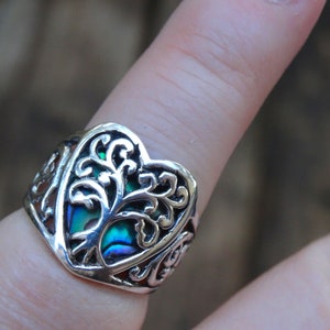 May include: A silver ring with a heart-shaped design featuring a tree of life motif. The tree is made of abalone shell with iridescent green, blue, and purple hues.