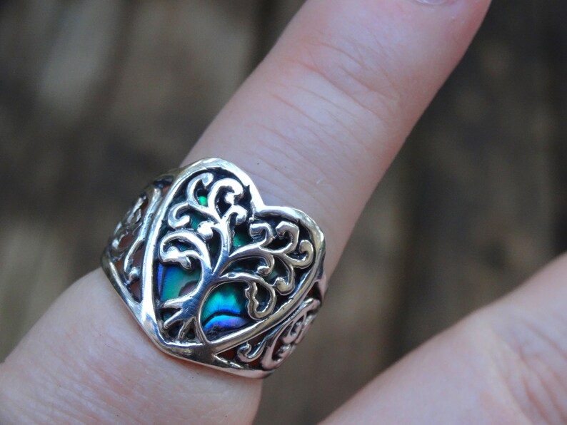 May include: A silver ring with a heart-shaped design featuring a tree of life motif. The tree is made of abalone shell with iridescent green, blue, and purple hues.