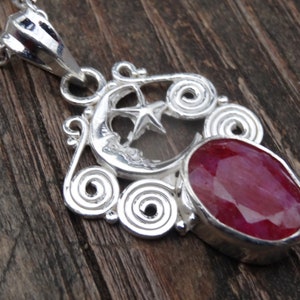 May include: A silver pendant necklace with a red gemstone and a crescent moon design. The pendant features a star inside the moon and intricate swirls around the gemstone.