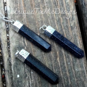 May include: Three dark blue gemstone pendants with silver caps. The pendants are shaped like long, thin rectangles with pointed ends. They are arranged on a wooden surface.