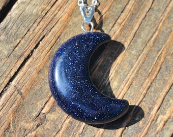 May include: A crescent moon pendant necklace. The pendant is a deep blue with sparkling inclusions, suspended from a silver chain. The necklace is set against a wooden background. The text "Unique To Me Design" is visible.