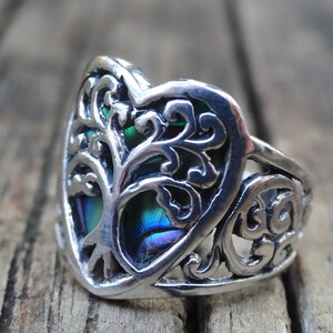 May include: A silver ring with a heart-shaped design featuring a tree of life with iridescent blue and green accents.