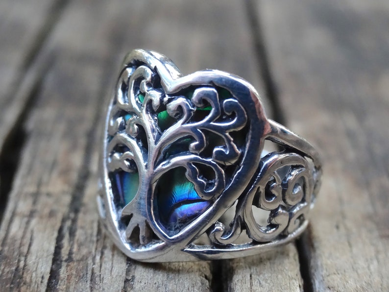 May include: A silver ring with a heart-shaped design featuring a tree of life with iridescent blue and green accents.