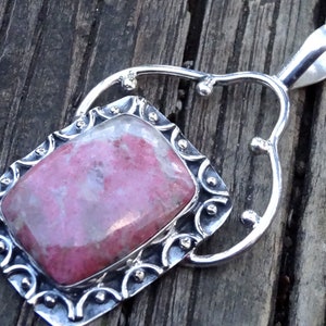 Peut inclure: Un collier pendentif en argent avec une pierre précieuse rose rectangulaire. La pierre est sertie dans un cadre argenté décoratif avec des détails ornés. Le pendentif est suspendu à une fine chaîne en argent, présentant un design unique et élégant.