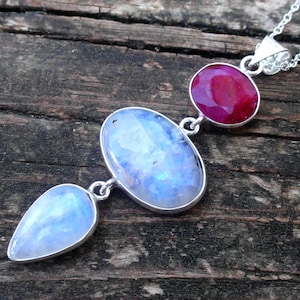 May include: A silver necklace featuring a large oval moonstone, a teardrop-shaped moonstone, and a ruby gemstone. The gemstones are set in silver and connected by a delicate chain. The necklace is displayed on a wooden surface.