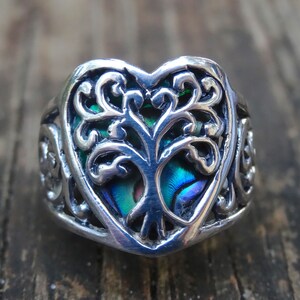 May include: A silver ring with a heart-shaped design featuring a tree of life motif. The tree is made of abalone shell with iridescent blue, green, and purple colors.