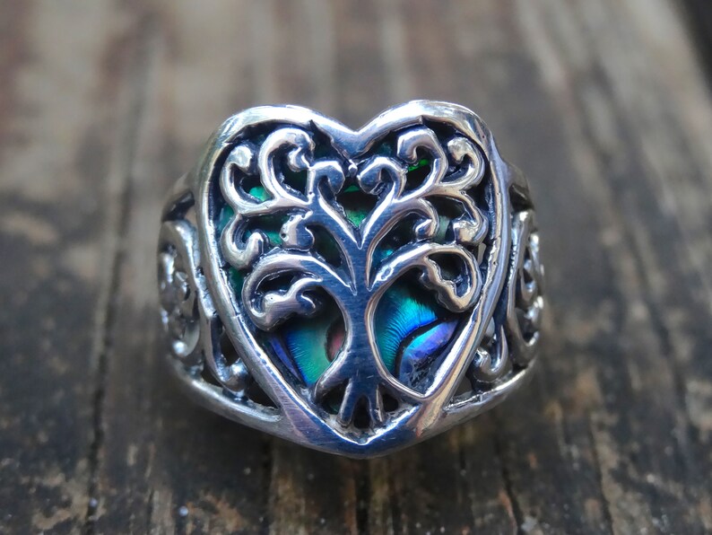 May include: A silver ring with a heart-shaped design featuring a tree of life motif. The tree is made of abalone shell with iridescent blue, green, and purple colors.
