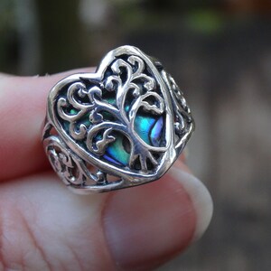 May include: A silver ring with a heart-shaped design featuring a tree of life motif. The tree is made of abalone shell with iridescent blue, green, and purple hues.
