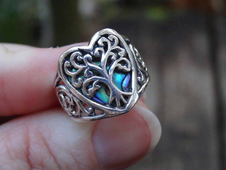May include: A silver ring with a heart-shaped design featuring a tree of life motif. The tree is made of abalone shell with iridescent blue, green, and purple hues.