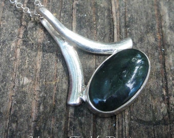 May include: A silver pendant with a dark green oval gemstone set in a geometric design. The pendant is on a silver chain.