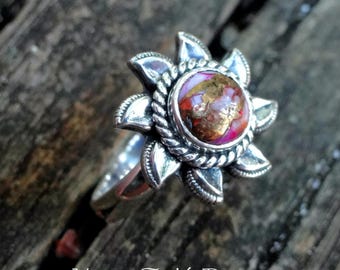 May include: A silver ring with a pink and gold gemstone set in a flower design. The ring is on a wooden surface.