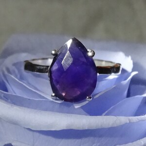 May include: A silver ring featuring a large, faceted amethyst gemstone in a teardrop shape. The ring is displayed against a blue background.