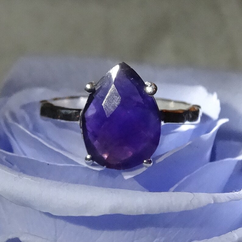 May include: A silver ring featuring a large, faceted amethyst gemstone in a teardrop shape. The ring is displayed against a blue background.
