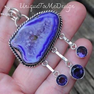 May include: A silver pendant necklace featuring a large purple agate stone with a crystalized centre. The pendant has three dangling charms with smaller purple amethyst stones.