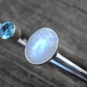 May include: A silver cuff bracelet featuring two gemstones. One is a small, round, light blue gemstone, and the other is a larger, oval, iridescent moonstone. The bracelet has a minimalist design.