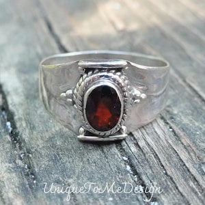 May include: A silver ring with a red gemstone set in a decorative bezel. The ring has a textured band and a simple design.