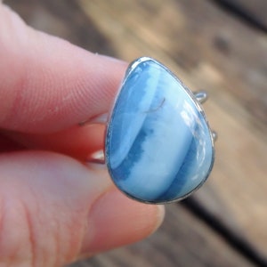 May include: A silver ring featuring a teardrop-shaped gemstone with a blue and white striped pattern. The ring's setting is silver, and the gemstone has a polished surface. The ring is shown against a blurred background.