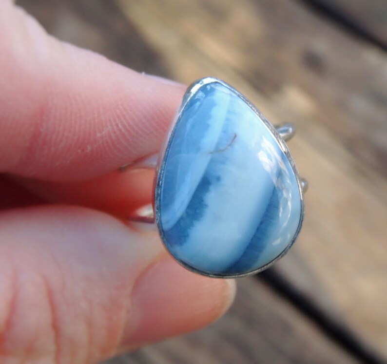May include: A silver ring featuring a teardrop-shaped gemstone with a blue and white striped pattern. The ring's setting is silver, and the gemstone has a polished surface. The ring is shown against a blurred background.