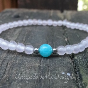 May include: A delicate bracelet made with light pink beads and a single turquoise bead in the center. The bracelet is accented with two silver beads on either side of the turquoise bead.