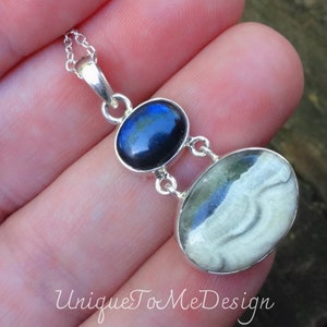 May include: A silver necklace with a pendant featuring two oval-shaped stones. The top stone is dark blue and the bottom stone is white with grey swirls. The necklace is on a hand.