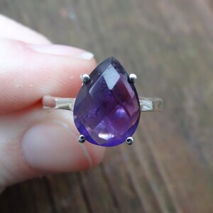 May include: A silver ring featuring a large, faceted amethyst gemstone in a pear shape.