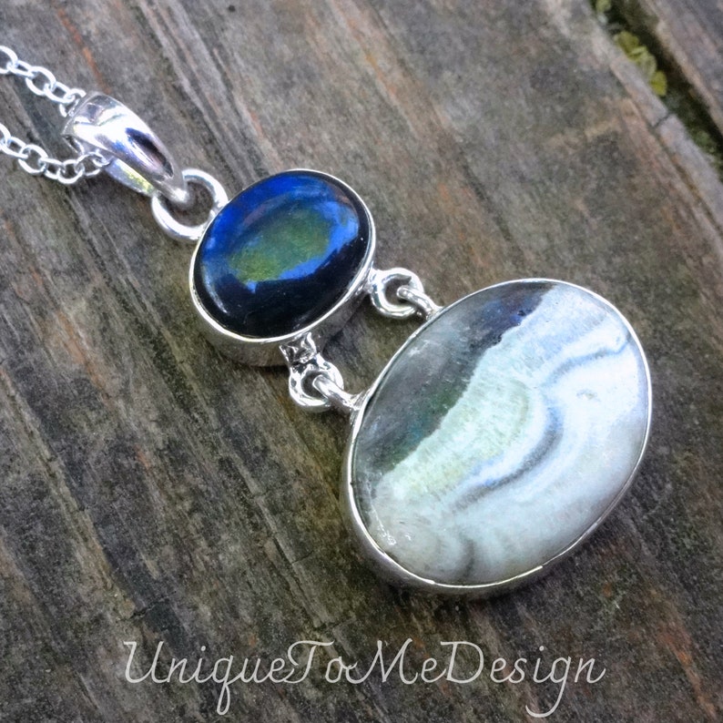 May include: A silver pendant necklace with two oval-shaped stones. The top stone is dark blue with green and gold iridescent accents. The bottom stone is white with grey and black swirls.