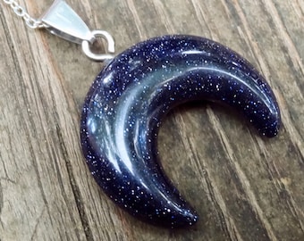 May include: A crescent moon-shaped pendant made of blue sandstone with silver flecks. The pendant is attached to a silver chain.