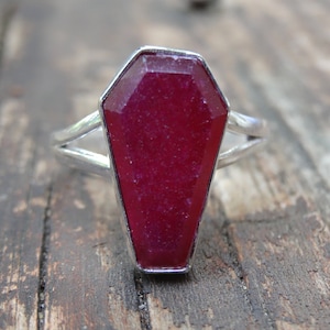 May include: A silver ring with a red gemstone shaped like a coffin.