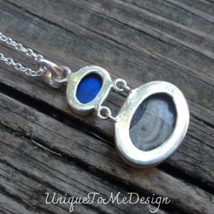 May include: A silver pendant necklace with two oval-shaped stones. The larger stone is grey with white swirls, and the smaller stone is blue. The pendant has a 925 stamp on the back.