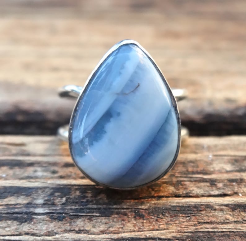 May include: A teardrop-shaped ring featuring a light blue gemstone with white and darker blue streaks. The stone is set in a silver-colored band. The ring is displayed on a wooden surface.