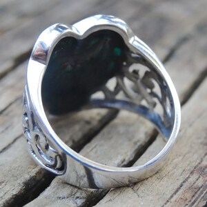 May include: Silver ring with a green stone set in a heart-shaped design. The ring has intricate filigree details around the stone.