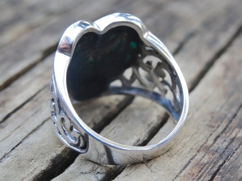 May include: Silver ring with a green stone set in a heart-shaped design. The ring has intricate filigree details around the stone.