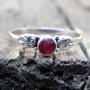 May include: A silver ring with a red gemstone set in a bezel setting. The ring has two decorative side pieces with a floral design.