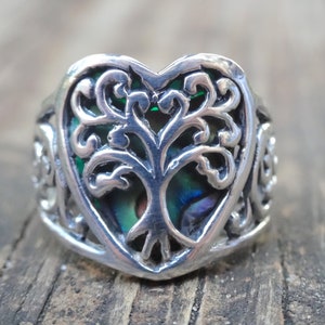 May include: A silver ring with a heart-shaped design featuring a tree of life motif. The tree is surrounded by a green abalone shell.