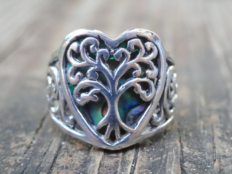 May include: A silver ring with a heart-shaped design featuring a tree of life motif. The tree is surrounded by a green abalone shell.