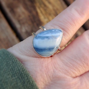 May include: A silver ring featuring a pear-shaped, blue and white gemstone. The stone has a layered appearance with bands of light and dark blue. The ring is set in a silver band with a simple, elegant design.