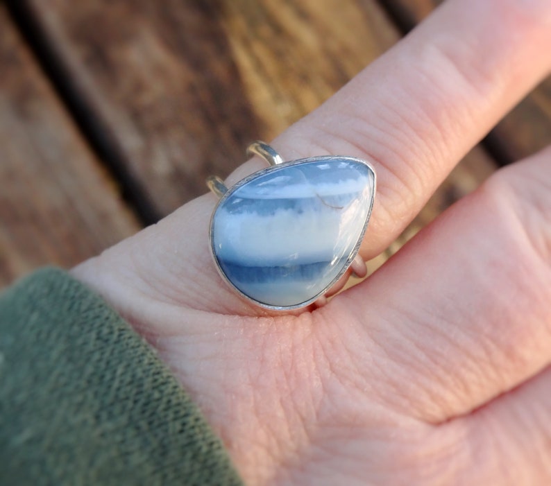 May include: A silver ring featuring a pear-shaped, blue and white gemstone. The stone has a layered appearance with bands of light and dark blue. The ring is set in a silver band with a simple, elegant design.
