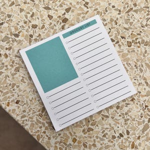 May include: A white grocery list notepad with a teal-colored block and the words "GROCERY LIST" printed at the top. The notepad has lines and circles for checklist items. The notepad is on a speckled surface.