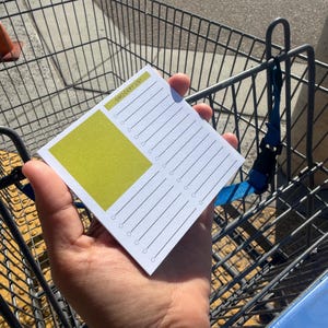 May include: A white notepad with a green square and the words "Grocery List" printed on it. The notepad has lines and circles for writing. The notepad is being held in front of a shopping cart.