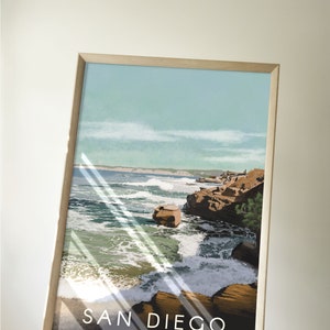 以下が含まれることがあります： サンディエゴ、カリフォルニアのビーチシーンを描いたヴィンテージな旅行イラストの額装ポスター。ポスターには青い空、白い雲、そして波が打ち寄せる岩だらけの海岸線が描かれている。ポスターの下部には黒い背景に白い文字で「SAN DIEGO - California -」と印刷されている。