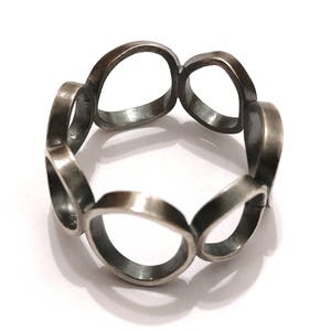 Può includere: Un anello d'argento con un design unico caratterizzato da più cerchi interconnessi. L'anello è realizzato in metallo liscio e lucido e presenta un'estetica moderna e minimalista.