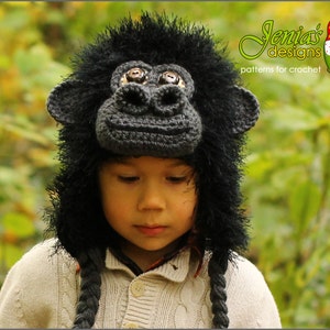 May include: A child wearing a black crocheted gorilla hat with brown eyes. The hat has a long tail that is braided. The child is wearing a light brown sweater.
