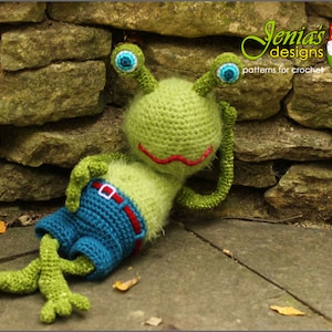 Könnte beinhalten: Ein grüner Häkel-Frosch mit blauen Augen und einem roten Mund, der blaue Jeans-Shorts mit einem roten Gürtel trägt. Der Frosch liegt auf der Seite mit ausgestreckten Beinen.
